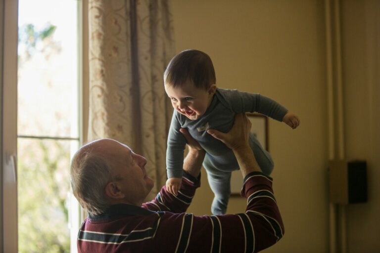 man wearing maroon, white, and blue stripe long-sleeved shirt lifting up baby wearing gray onesie